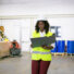 Focused female logistic employee in hardhat and safety vest Focused female logistic employee in hardhat and safety vest walking in warehouse, carrying open folder, looking through document. Copy space, front view. Labor and inspection concept